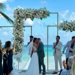 A bride and groom kissing under a white floral arch on a beach wedding ceremony setup.