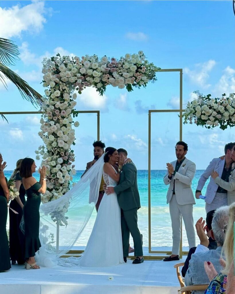 A bride and groom kissing under a white floral arch on a beach wedding ceremony setup.