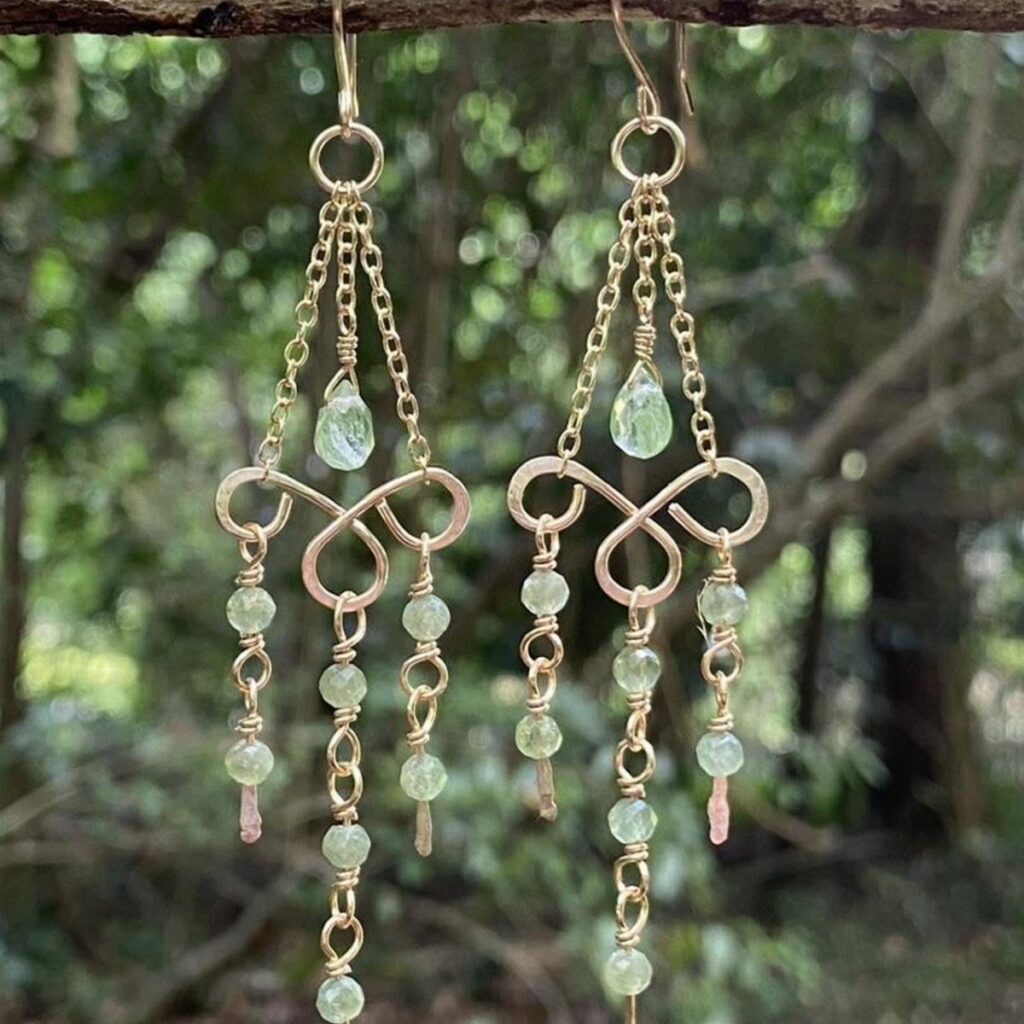 Gold chandelier earrings with light green gemstones hanging against a blurred green forest background.