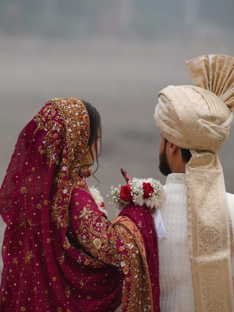 A bride in maroon and gold attire faces a groom wearing a cream turban and white sherwani.