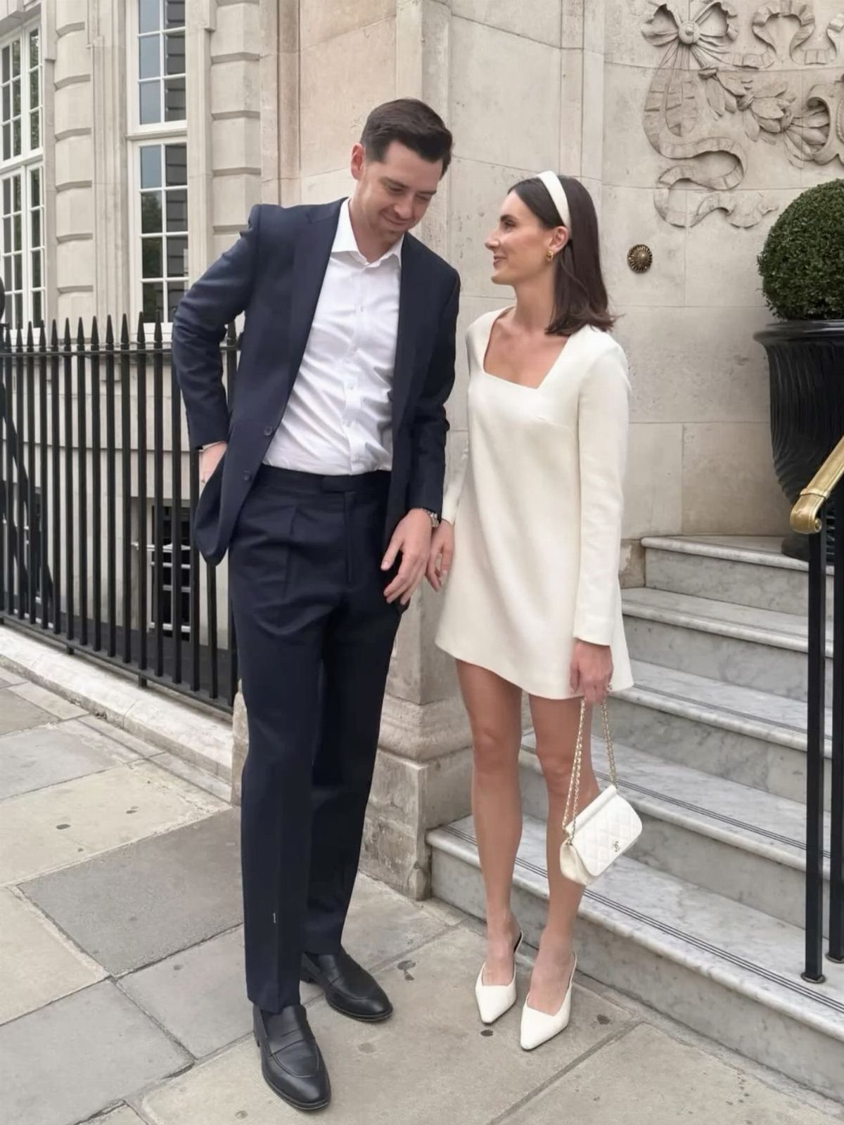 A woman in a short white crepe dress and a man in a navy suit standing on stone steps.