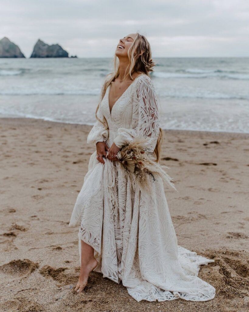 A woman in a white lace flowing dress stands on a beach holding a bouquet.
