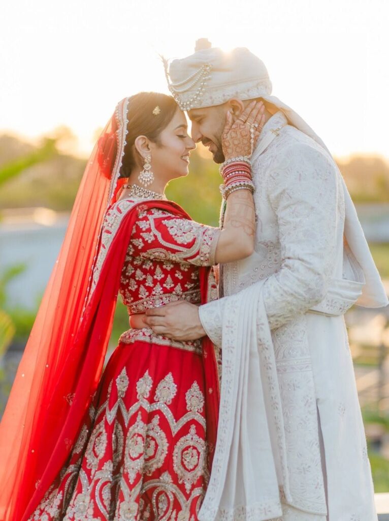 A bride and groom in traditional Indian wedding attire touching foreheads outdoors during sunset.