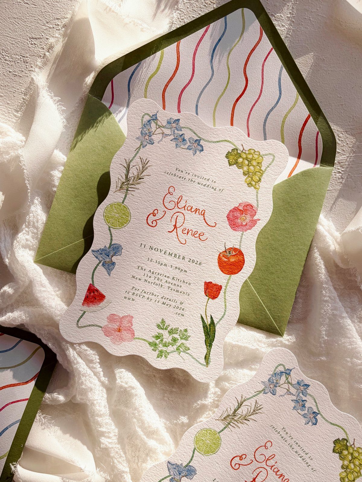 Botanical wedding invitation with colorful floral illustrations resting inside a green lined envelope on white fabric.