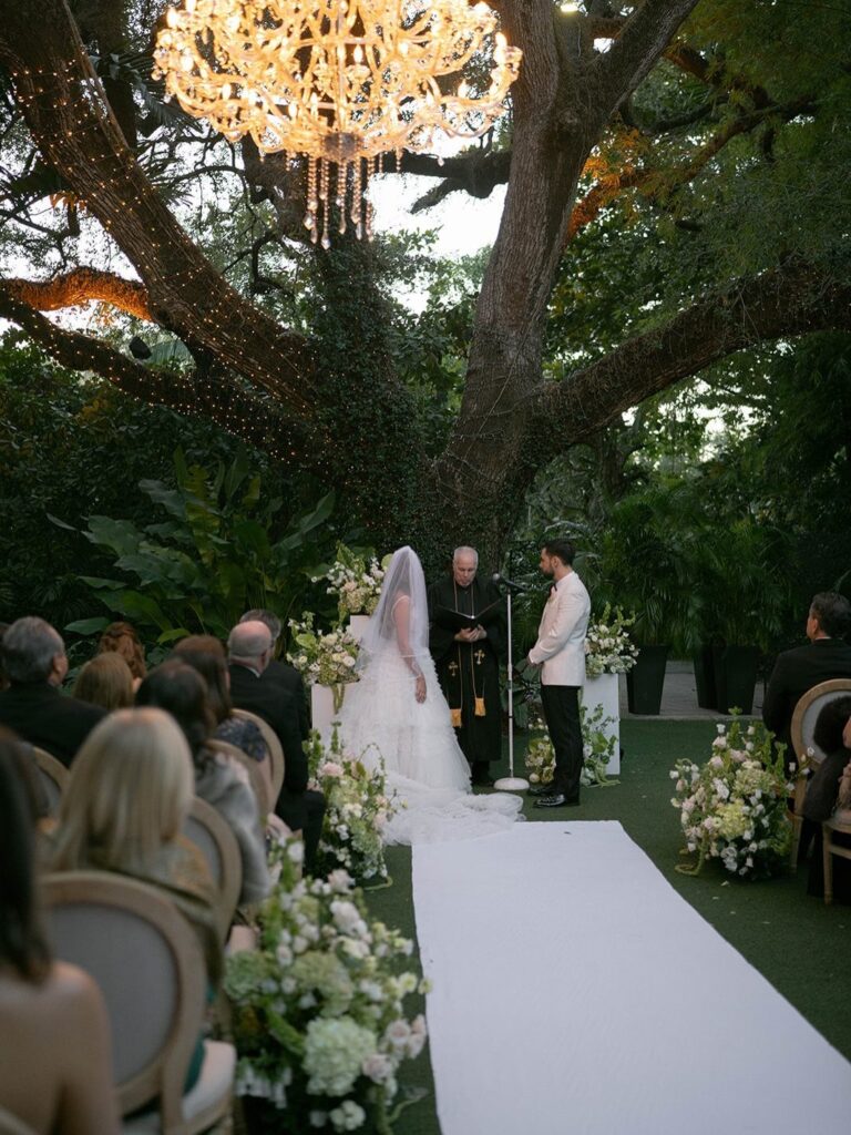 A bride and groom exchange vows during a garden wedding ceremony under a large tree.