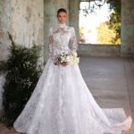 A woman wearing a long-sleeved white lace wedding gown holding a white flower bouquet.