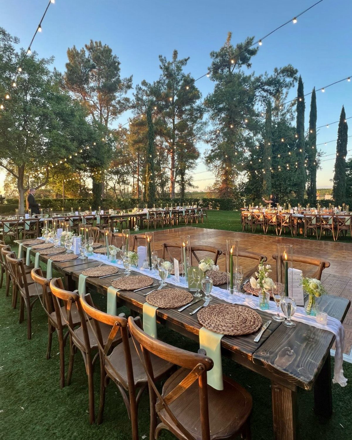 A long rustic wooden dining table set with woven placemats and flowers under outdoor string lights.