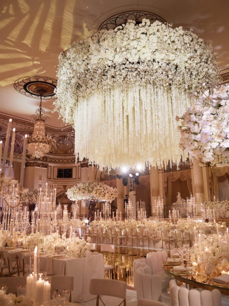Large white floral chandeliers hanging over elegantly set wedding reception tables in a grand ballroom.