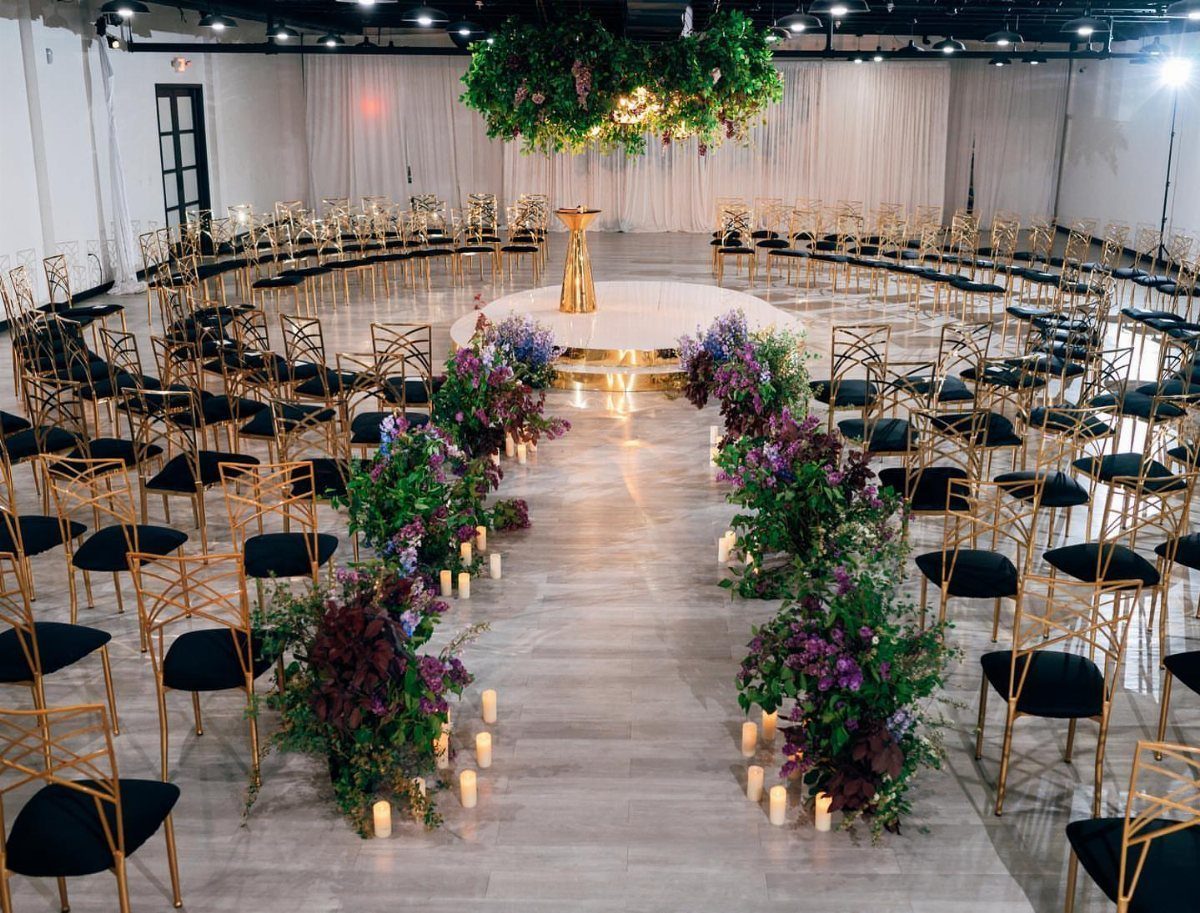 Gold chairs arranged in a circle around a white platform with a floral aisle and greenery.