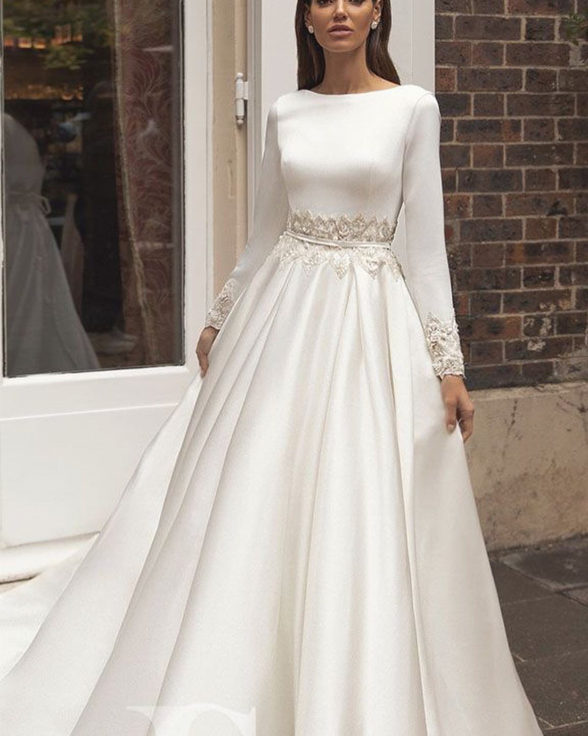 A woman in a white long-sleeved wedding dress with lace detailing stands near a brick wall.