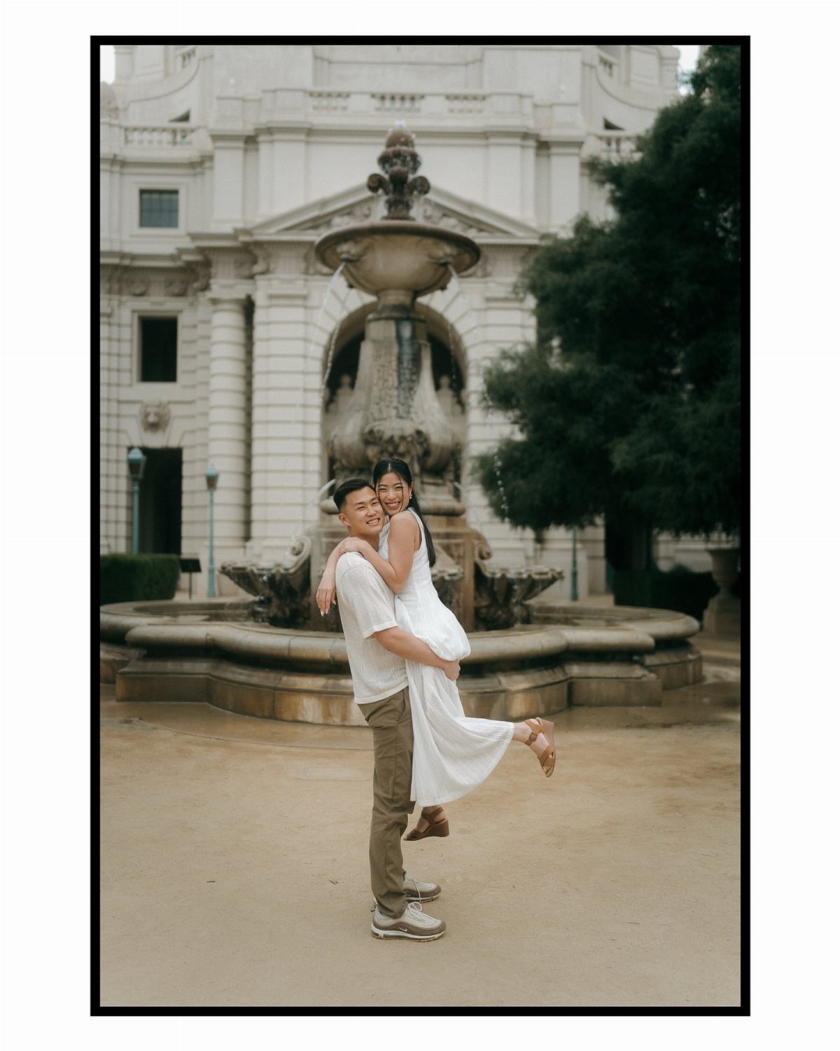 A happy couple embracing and smiling in front of a large stone fountain in a city plaza.
