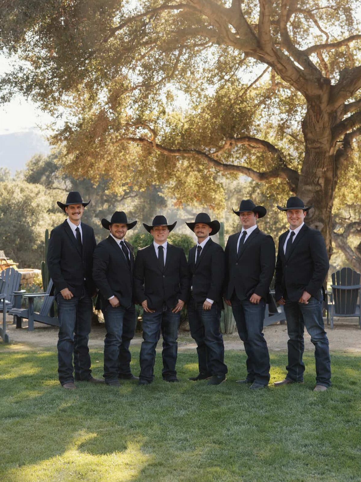 Six men wearing black suit jackets, black ties, jeans, and cowboy hats standing outdoors.