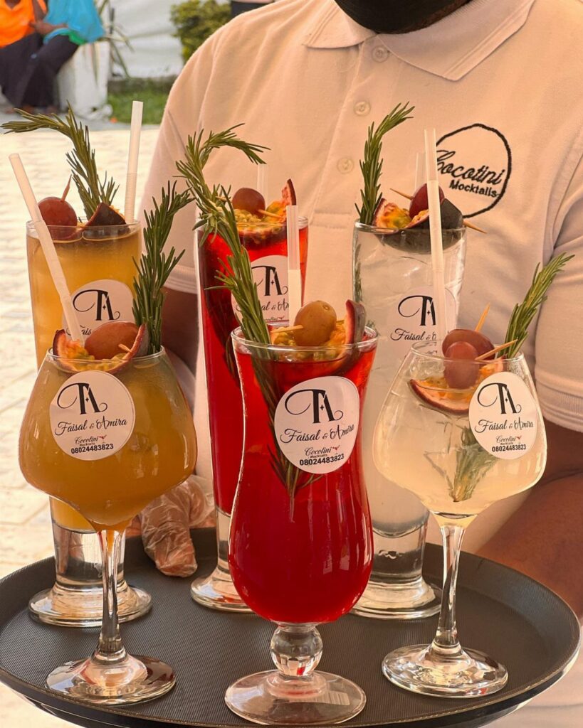 Four colorful garnished cocktails on a tray held by a person wearing a white polo shirt.