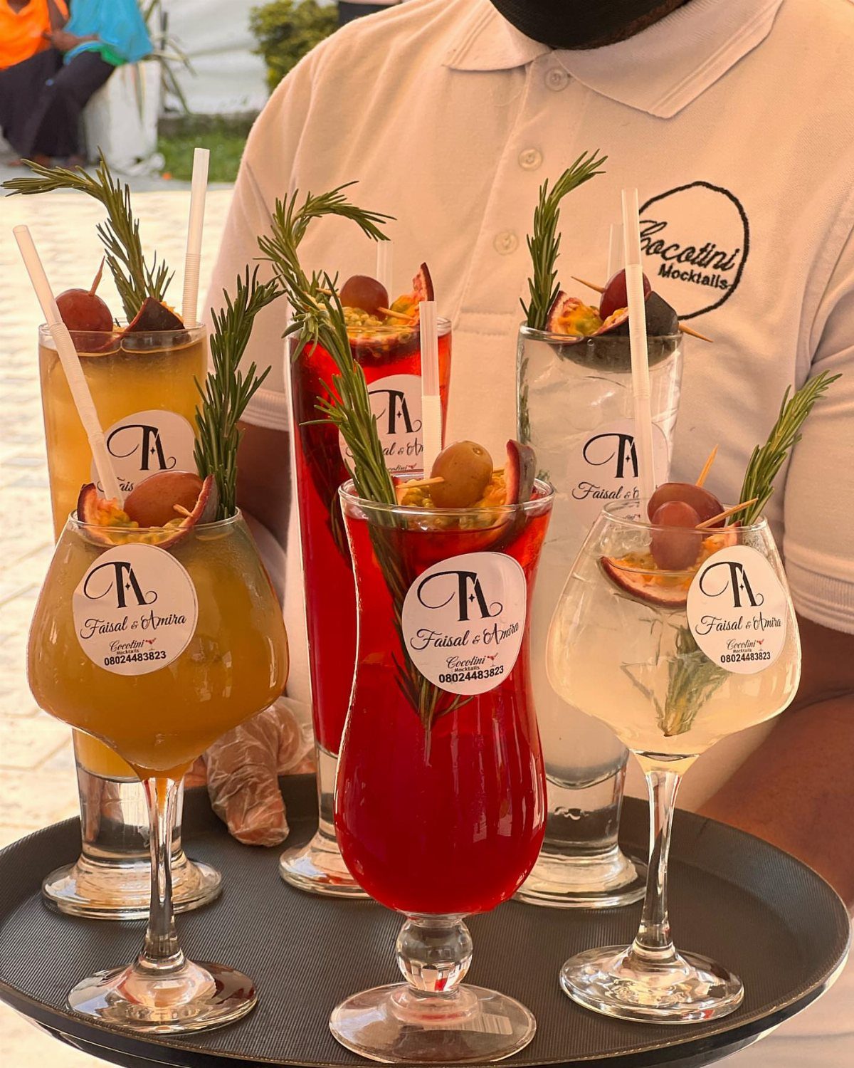 Four colorful garnished cocktails on a tray held by a person wearing a white polo shirt.
