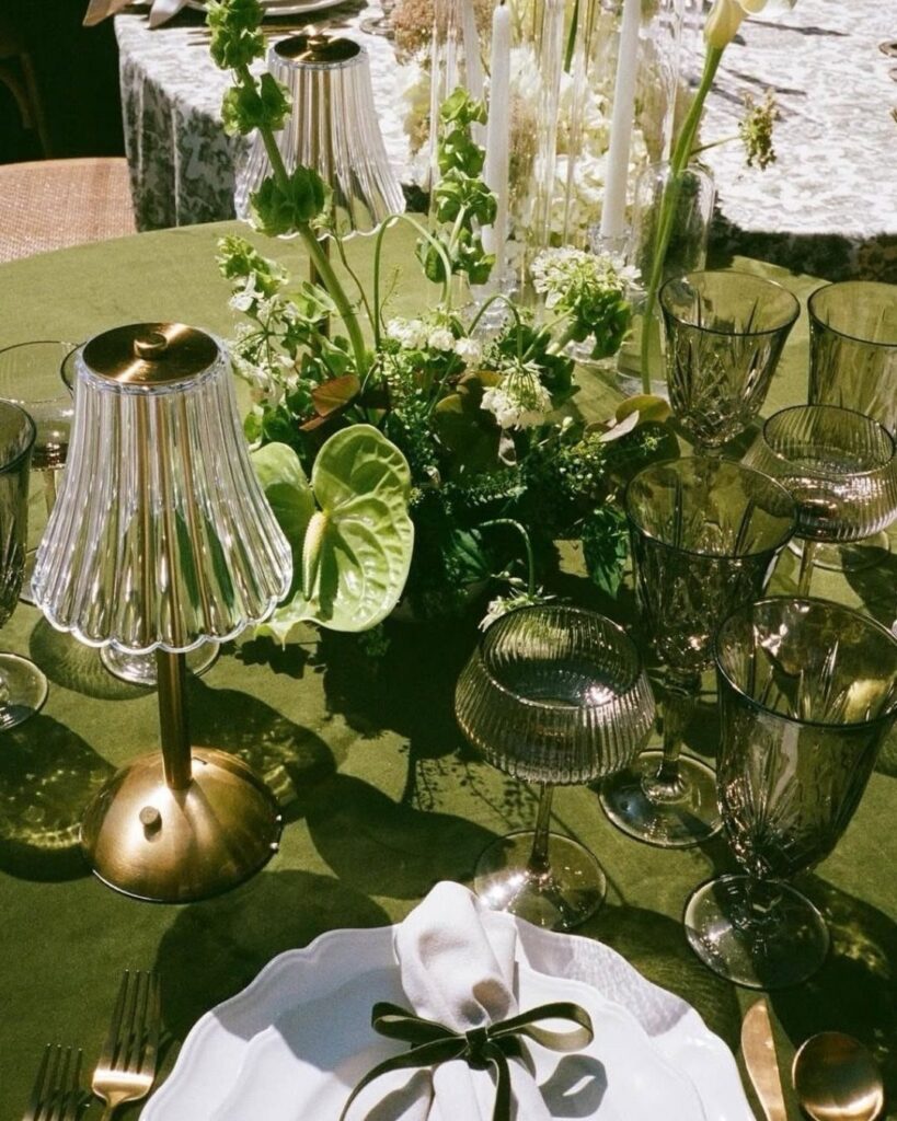 Green wedding table setting with floral centerpieces, glassware, and gold accents on a green tablecloth.
