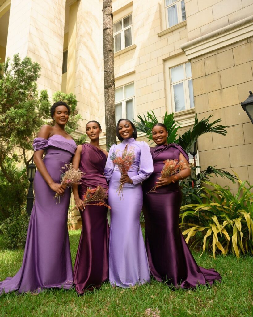 Four women in violet and purple satin bridesmaid dresses holding small floral bouquets outdoors.
