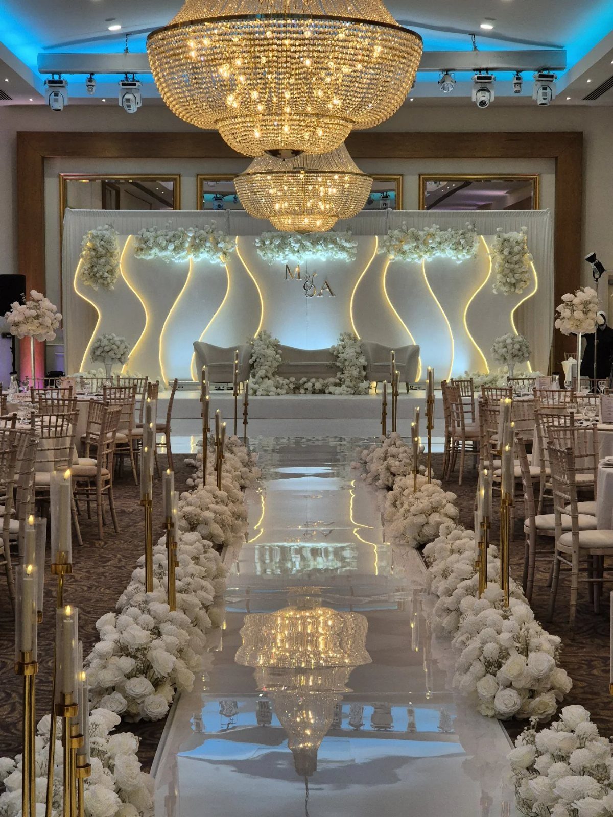 White floral arrangements line a reflective aisle runner leading to a decorated wedding stage with gold accents.